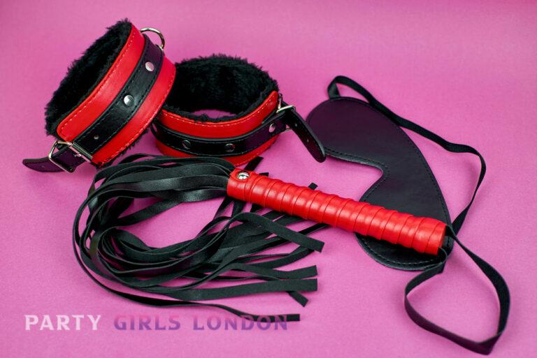 Red and black leather cuffs, blindfold, and soft whip arranged on pink background symbolising playful control and trust in London escorts’ fantasy experiences