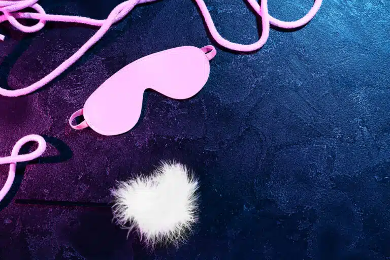 Pink blindfold, soft white feather, and rope arranged on dark textured background under neon light