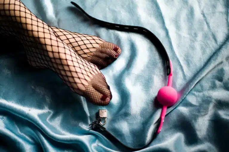 Close-up of a Knightsbridge escorts feet in fishnet tights resting on blue satin sheets beside a pink accessory, suggesting playful elegance and intrigue
