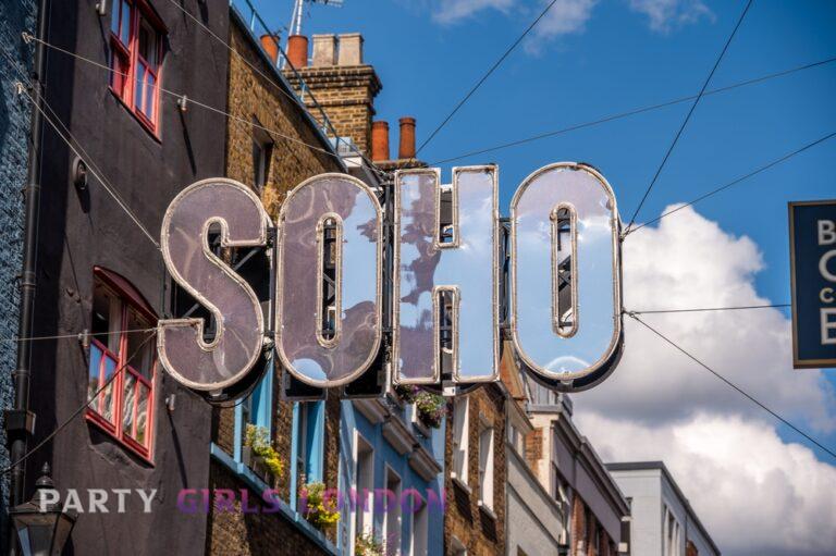 large mirrored Soho sign hanging above colourful London buildings under a blue sky, symbolising one of the city’s most iconic nightlife districts