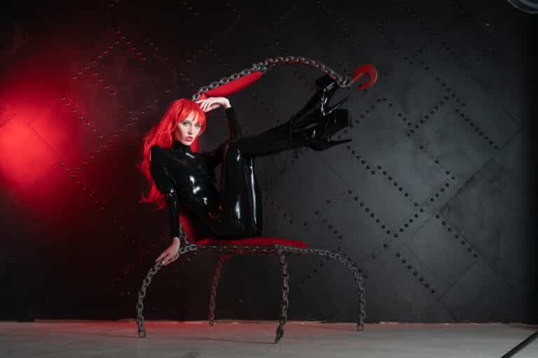 Marylebone dominatio n escort in a black latex catsuit with red hair posing on a metal chair under red light in a studio
