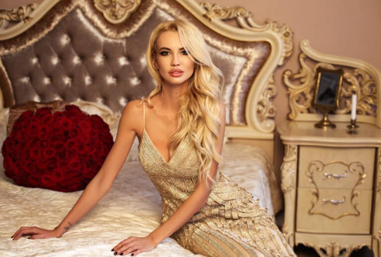 Blonde high class Kensington escort in a golden evening gown sitting gracefully on a luxury bed in London
