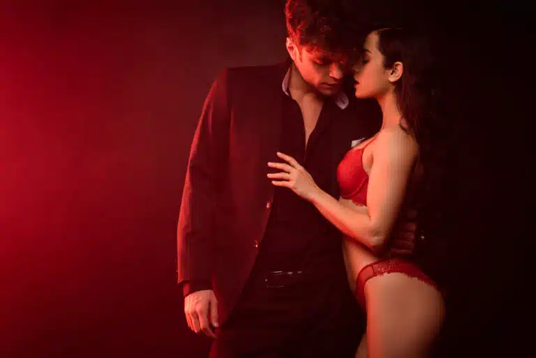 Woman in red lingerie and man in a dark suit sharing an intimate, close moment under warm red lighting, suggesting curiosity and emotional connection